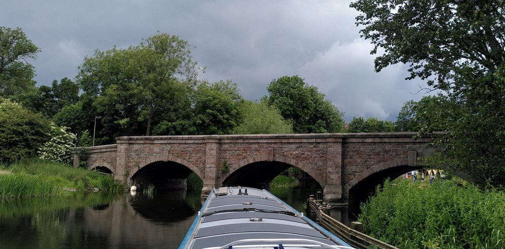 Centre arch.  From the stern deck of a narrowboat the centre arch of a stone road bridge is directly ahead.  A metal guard rail has been placed to prevent boats being taken under the arch to the right.  There is a very dark cloud in the sky above.