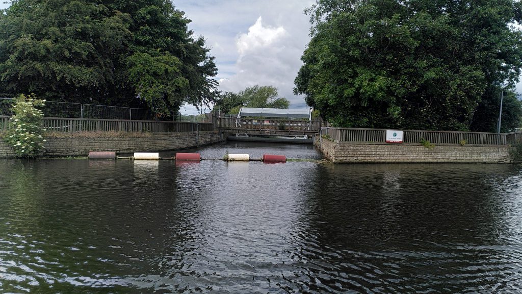 Flood channel.  A side channel from a canal blocked off with red and white floats.  A short distance down the channel is a rotating barrier - currently closed.