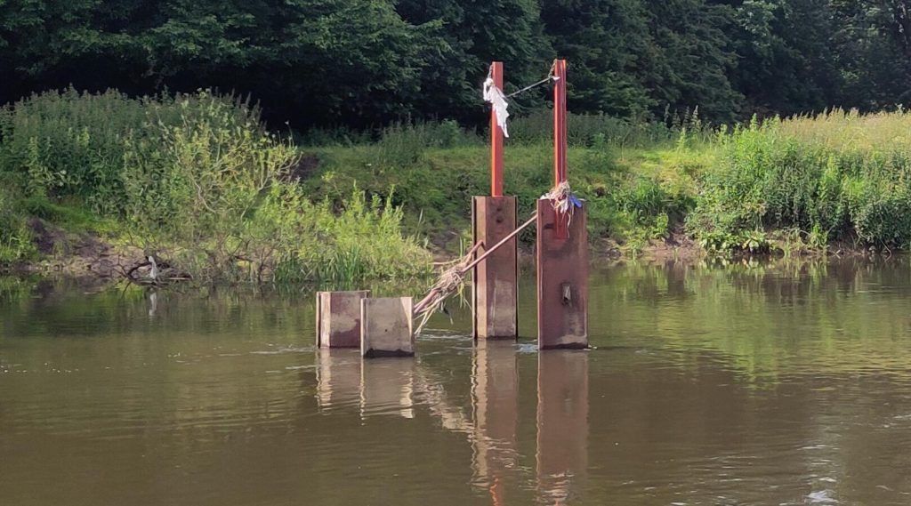 Hazard marker.  Four lengths of piling protruding above the water of the river.  Two have extra lengths of metal welded on that are higher than the bank.