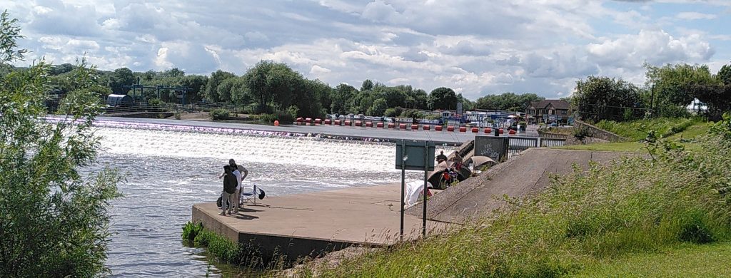 Beeston Weir.  The water of the river cascades down the steeped weir.  Above it a line of bright orange floats mark the danger for boats.  The upstream far bank beyond is lined with moored boats.