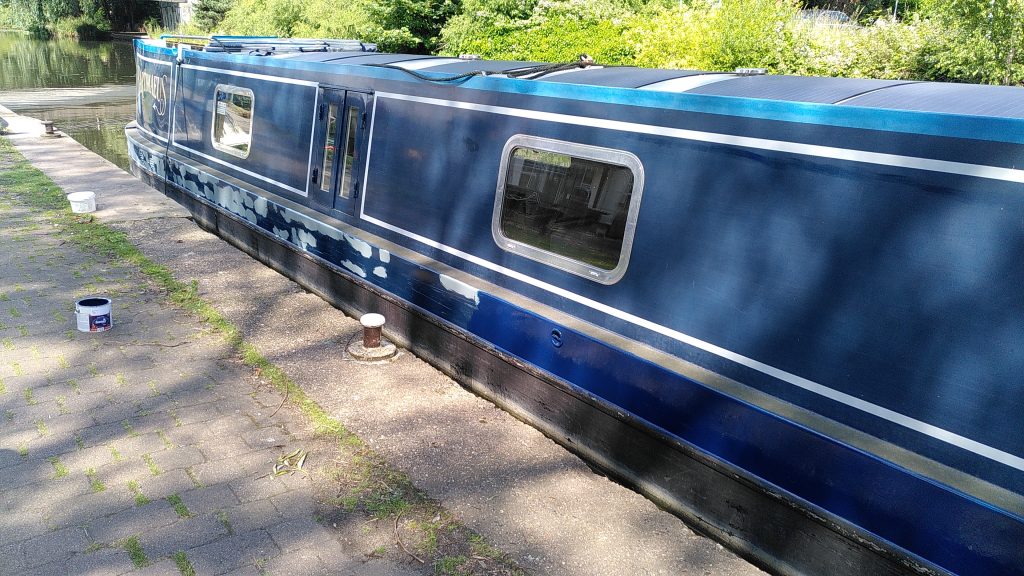 Partly done. A narrowboat is moored against a low concrete bank.  The paintwork below the gunwale is a patchwork of grey undercoat at the stern of the boat.  Around the middle of the boat the paint below the gunwale is a uniform dark blue.