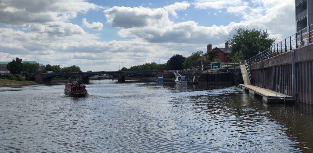 Up river.  Looking back up the River Trent.  On one bank there is a jetty for the lock.  Moored boats sit at another jetty upstream.  A three arch bridge crosses the river further up.  A narrowboat driving upstream seems very small in the wide river.  
