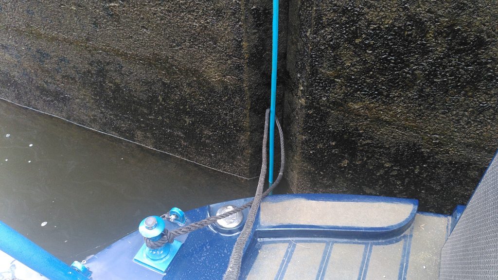Control wire.  The stern deck of a narrowboat is resting close to a lock wall.  A rope tied to a dolly on deck is wrapped around a vertical cable covered in blu plastic which is recessed in to the lock wall.