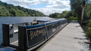 Sunny mooring. A narrowboat is moored on to a floating pontoon at the side of a river. The tree-lined banks of the river extend to the horizon. There are light clouds in the sky above.