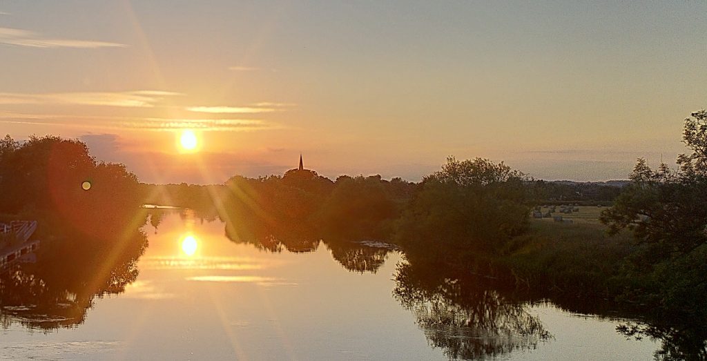 Sunset.  The sun is low in the sky and reflected in the wide river.  The skyline is mostly trees but with a church spire prominent.  Some low clouds glow iwith the same golden light as the sun.