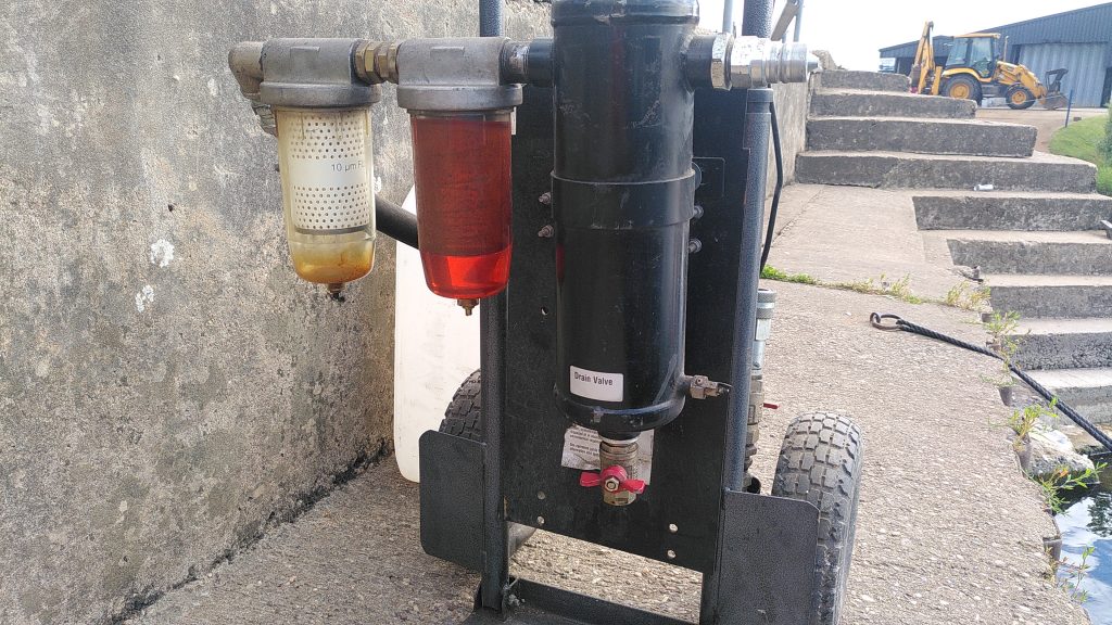 Fuel polisher. Some machinery is mounted on a two wheeled trolley. Three cylinders are arranged in a line. The first is large and black. The second is transparent and filled with red diesel. The third has a white filter but is otherwise empty. The kit is sitting on a narrow concrete path,