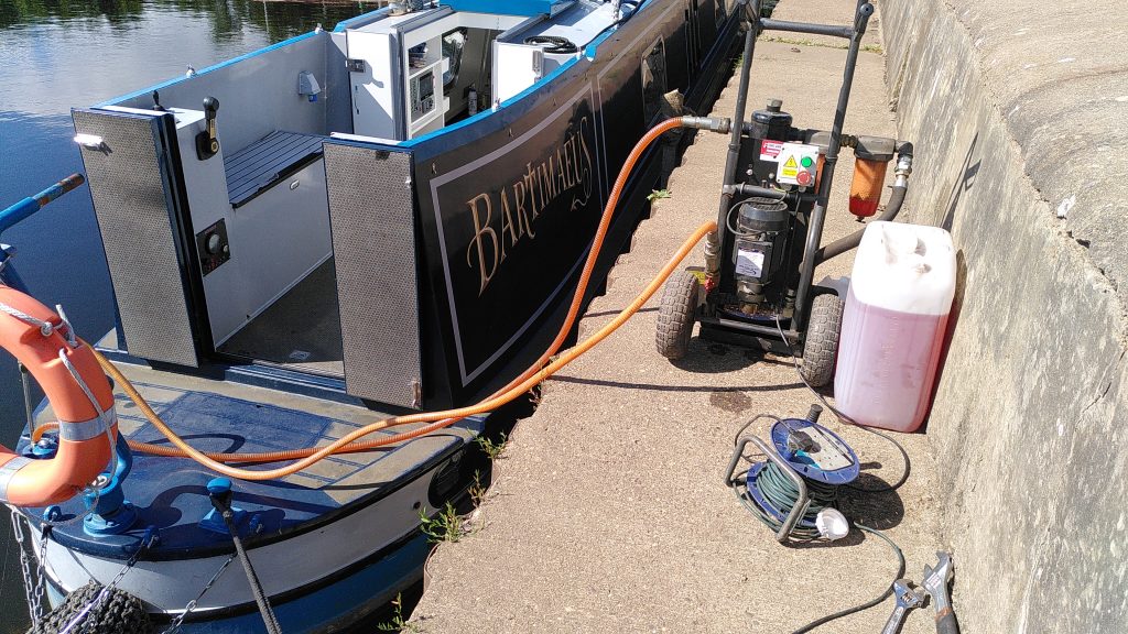 Polishing. Hoses run from a fuel polisher on to the deck of a moored narrowboat. Next to the machine is a large container containing about 30 litres of red diesel.
