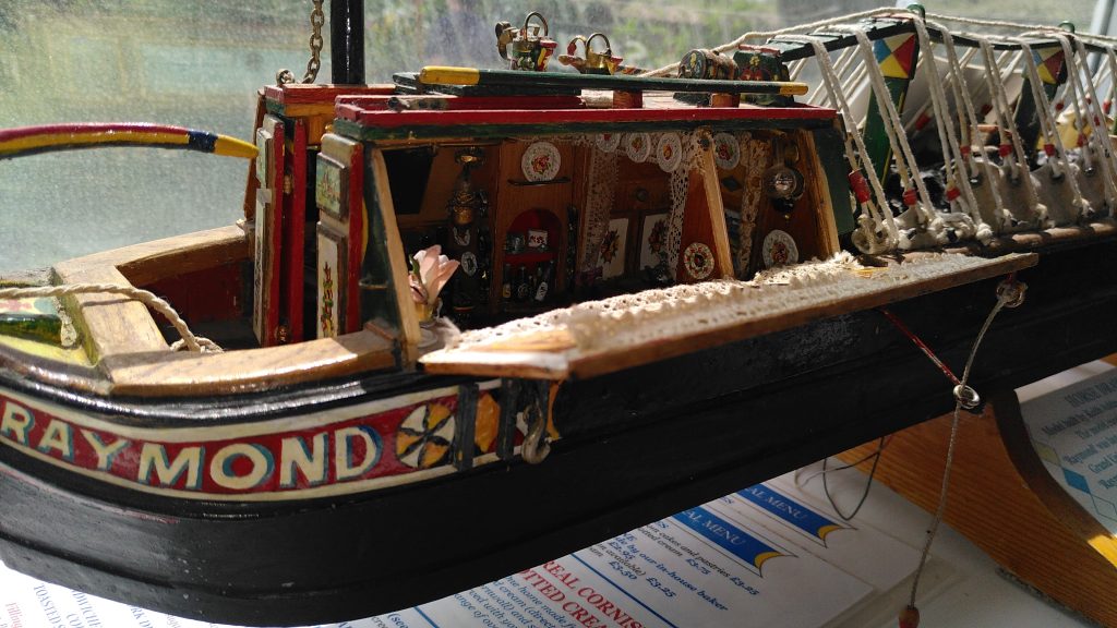 Raymond.  A model of a narrowboat called Raymond sits in the window of a tea shop.  The wall of the butty - the living quarters at the stern of the boat - has been hinged down to reveal the detail inside.