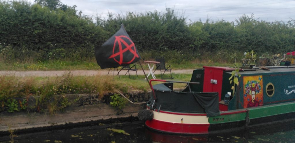 Anarchy flag.  A large black flag with a red capital A in a circle is flying from the tiller of a narrowboat.  The flag is as high as the cabin sides.