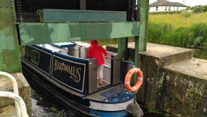 Guillotine lock. The stern of a narrowboat is passing under the guillotine gate at one end of a lock. The driver is ducking to get under the gate and counterweight and has her hood up to avoid the drips.