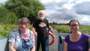 Gail, Rab and Clare. A man is at the tiller of a narrowboat. Two women are sitting on the bench seats either side of the boat. The canal behind is lined with vegetation.